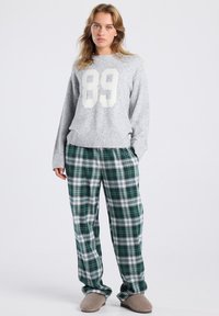 Femme portant un pull gris avec le chiffre "89", un pantalon à carreaux vert et blanc, et des pantoufles beiges, se tenant les mains dans les poches sur un fond blanc.