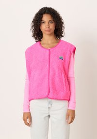 Flis brezrokavnik v svetlo roza barvi z mehko, teksturirano površino. Ima okroglo izrez, pritisne gumbe in majhno emblem na prsih.