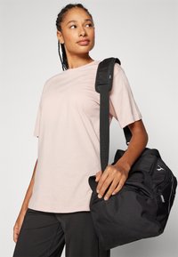 Sort sportstaske med tekstureret stof, dobbelte håndtag og justerbar rem, parret med en lyserød t-shirt med korte ærmer og sorte bukser.