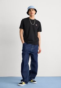 Black short-sleeve t-shirt with small logo, blue bucket hat, loose-fit dark blue jeans, and black sneakers with yellow accents.