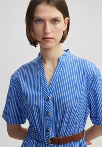 Abito camicia a righe blu e bianche con maniche corte, colletto alla coreana, bottone frontale e una cintura marrone in vita. Tessuto dalla texture liscia.