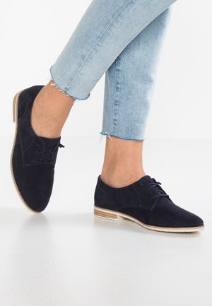 Chaussures oxfords en daim bleu marine avec un bout arrondi, un design à lacets et une semelle en bois clair. Portées avec un jean bleu clair retroussé.