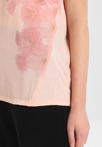 Blouse rose clair avec un imprimé floral sur le côté gauche, fabriquée dans un tissu lisse et transparent, avec une coupe décontractée et un ourlet arrondi.