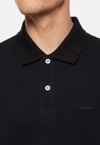 Detailný záber na osobu v čiernom polo tričku s dvoma bielymi gombíkmi a malým čiernym vyšitým logom na hrudi.