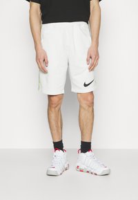 Pantaloni sportivi bianchi con logo Nike nero, caratterizzati da accenti laterali verdi. Indossati con scarpe bianche con dettagli rossi e calze nere.