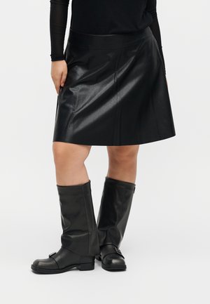 Persona que lleva una falda de cuero negra con botas negras hasta la rodilla y una camiseta negra de manga larga sobre un fondo liso.