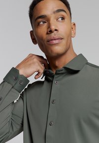 Groene langekous shirt met een ontspannen kraag, voorzien van donkere knopen en een soepele textuur. Model raakt de kraag aan, waardoor de pasvorm wordt getoond.