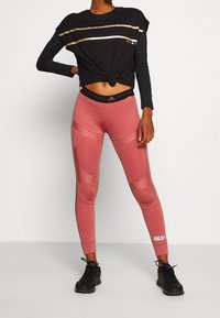 Top negro de manga larga con cintura anudada y rayas doradas. Leggings rosas con paneles transparentes y cintura negra con logo. Calzado negro.