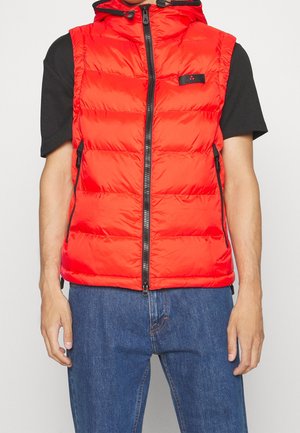 Homme portant un gilet matelassé sans manches rouge vif avec une fermeture éclair à l'avant et une capuche, par-dessus un t-shirt noir à manches courtes et un jean bleu.