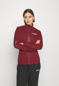 Bordeauxfarbene Softshelljacke mit hohem Kragen, Reißverschlusstaschen und schwarzen Akzenten. Sie verfügt über ein tailliertes Design und eine glatte Textur. Logo auf der Brust angebracht.