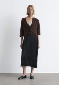 Brauner, flauschiger, kurzärmeliger Cardigan über einem schwarzen Satin-Kleid mit Spitze. Schwarze flache Schuhe runden den Look ab. Einfaches und elegantes Design.