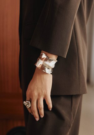Hand met een bruin pak, versierd met een brede, gesmede zilveren armband en een chunky, abstracte zilveren ring om de wijsvinger.