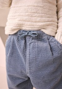 Gros plan d'une personne portant un pull en maille beige clair rentré dans un pantalon en velours côtelé bleu avec une taille élastique et un lien devant.