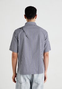 Mand med kort, krøllet hår iført en sort-hvid ternet kortærmet skjorte og lyse denimshorts, stående med ryggen til kameraet.