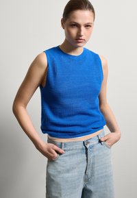 Blaues ärmelloses Stricktop mit strukturiertem Muster, Rundhalsausschnitt und lockerer Passform. Kombiniert mit hellblauen Jeans.