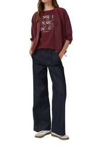 Burgunderfarbene Zip-Jacke über einem Grafik-T-Shirt, kombiniert mit dunklen weit geschnittenen Jeans und beigen Sneakern. Besitzt eine lockere Passform und ein lässiges Design.