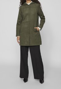 Olivgrön kappa med huva, tillverkad av mjukt tyg, har rak skärning, långa ärmar och två sidofickor. Bärs över svarta vida byxor.