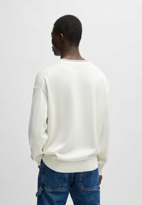 Maglione bianco lavorato a maglia con collo rotondo, polsini e orlo a coste, e dettagli testurizzati sottili vicino al collo, indossato con jeans di denim blu.