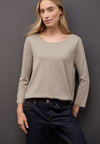 Hellbeige Langarmshirt mit rundem Ausschnitt, kombiniert mit dunklen Denim-Jeans mit orangen Nähten und Fronttaschen. Glatte Textur.