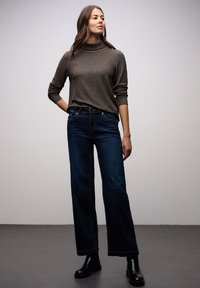 Maglione marrone a collo alto con maniche lunghe abbinato a jeans a gamba larga blu scuro. Stivali neri alla caviglia visibili; dettagli di cucitura sottili sui jeans.