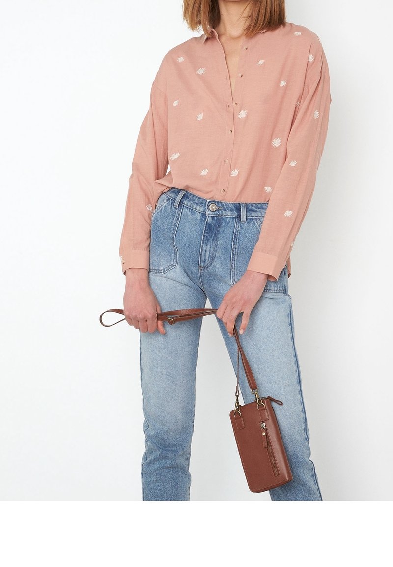 Femme portant une chemise rose à boutons avec des imprimés blancs, un jean bleu taille haute, tenant une pochette marron par sa sangle.