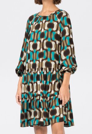 Femme portant une robe ample à manches longues avec une jupe à volants, arborant un motif géométrique marron, turquoise et crème.