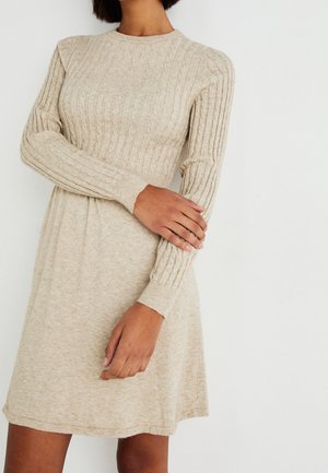 Femme portant une robe beige en maille côtelée à manches longues, debout avec un bras croisé et l'autre main reposant sur sa manche.