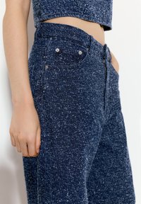 Kalhoty připomínající džíny v tmavě modré barvě s texturovaným povrchem. Mají vysoký pas, přední kapsy a detaily ze stříbrného kovu.