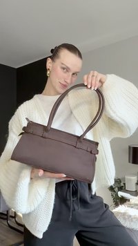 Mala de mão de couro castanho com formato estruturado, alças duplas e costura decorativa. Usada com um cardigan branco de malha e calças pretas.