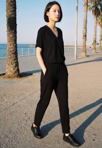 Mlada žena u crnoj odjeći hoda sunčanom šetnicom uz more obrubljenom palmama i ogradom uz vodu.