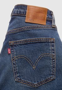 Poche arrière et ceinture d’un jean en denim bleu Levi’s 501 montrant un patch en cuir de la marque et une étiquette en tissu rouge Levi’s.