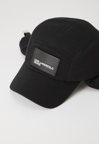 Čierna čiapka vyrobená z mäkkej tkaniny, s pevnou konštrukciou, prešitými detailmi a matnou čiernou koženou nášivkou s nápisom „Karl Lagerfeld Jeans.“