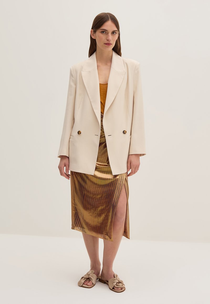 Blazer doppiopetto crema, gonna setosa a strisce oro e marrone con spacco laterale e sandali beige annodati. Texture morbide e vestibilità rilassata.