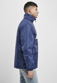 Tamsiai mėlynas užtrauktuku užsegamas švarkas su aukštu apykakle, elastinėmis rankogaliais ir šoniniais kišenėmis. Pagamintas iš blizgaus, lygio medžiagos, su baltu logotipu ant priekio.