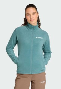 Giacca in pile di colore teal, con colletto alto e zip frontale. Logo sul petto. Tessuto morbido con maniche lunghe e tasche laterali.