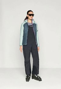 Woman wearing teal and navy ski jacket, black ski overalls, black ski boots, and large black sunglasses against plain background.