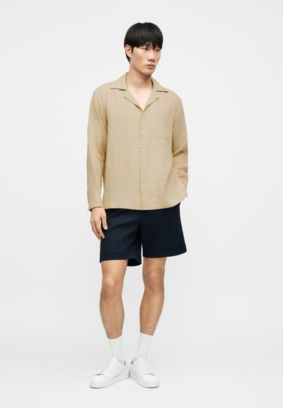 Chemise en lin beige avec col, manches longues, poche poitrine, associée à un short bleu marine et des baskets blanches. Design simple et couleurs neutres.