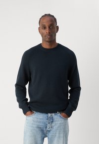 Maglione blu navy con collo a giro, polsini e orlo a coste. Texture morbida, realizzato in tessuto lavorato a maglia. Indossato sopra jeans azzurri.