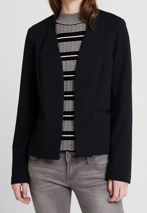 Nainen pukeutuneena mustaan bleiseriin mustavalkoraidallisen poolopaidan ja harmaiden farkkujen päällä, seisoo yksivärisen taustan edessä.