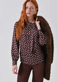 Giovane donna con lunghi capelli rossi, che indossa una blusa marrone a pois e pantaloni, tenendo un cappotto di pelliccia marrone sulla spalla.