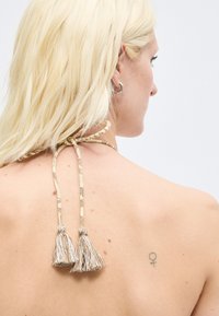 Femme blonde portant un top dos nu noué au cou avec des pampilles, montrant un petit tatouage du symbole féminin sur le haut de son dos.