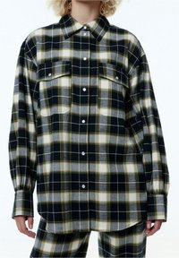 Karierter Button-up-Hemd in Schwarz, Grau und Gelb mit lockerer Passform, zwei Brusttaschen und abgerundetem Saum. Hergestellt aus weichem, wollähnlichem Material.