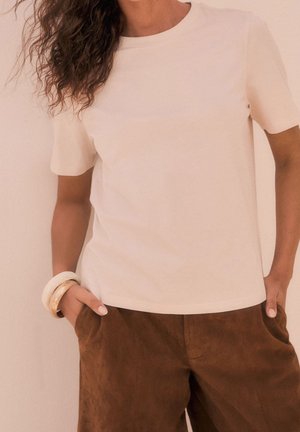 Vrouw draagt een effen beige top met korte mouwen en bruine suède broek, met handen in de zakken en armbanden om de linkerpols.