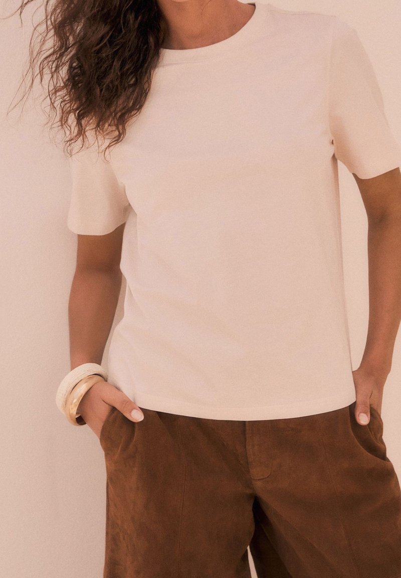 Femme portant un haut beige uni à manches courtes et un pantalon en daim marron, les mains dans les poches et des bracelets au poignet gauche.
