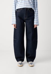 Jeans in denim scuro a gamba larga con cuciture visibili e tasche frontali, abbinati a una camicia a maniche lunghe a righe blu chiaro e sneakers argentate.