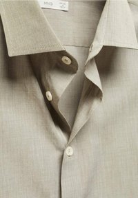 Chemise beige clair à boutons, fabriquée en tissu texturé, avec un col pointu et deux boutons blancs sur la patte de boutonnage.