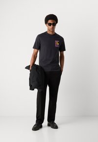 Sort bomulds t-shirt med et farverigt grafisk print på brystet. Modellen kombinerer den med sorte bukser og en sort jakke.