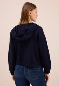 Femme aux cheveux mi-longs portant un sweatshirt à capuche bleu marine et un jeans en denim bleu, tournée de côté sur un fond beige.