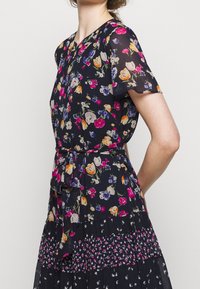 Blommig klänning med korta volangärmar, marinblå bas och flerfärgade blommönster. Knutet midjedetalj och lager-på-lager kjol med varierande blomstertryck.