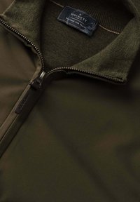 Tmavozelená bunda s rebrovaným golierom, čiastočne rozopnutý zips a štítok s nápisom „Hackett London Tailored for Travel“ vo vnútri výstrihu.